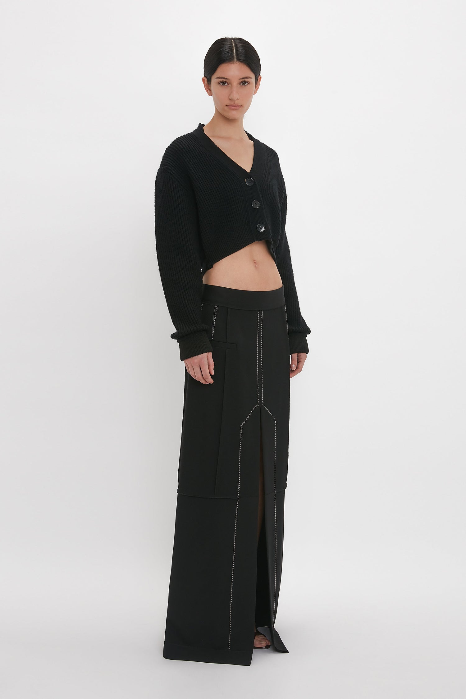 A person stands against a white background wearing a black cropped cardigan and a Victoria Beckham Deconstructed Floor-Length Skirt In Black.