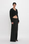 A person is standing against a white background, wearing a black Cropped V-Neck Cardigan In Black and a black high-waisted long skirt with front slits. The wardrobe’s chic simplicity is elevated by the compact ribbed knit of the cardigan, providing both texture and style.