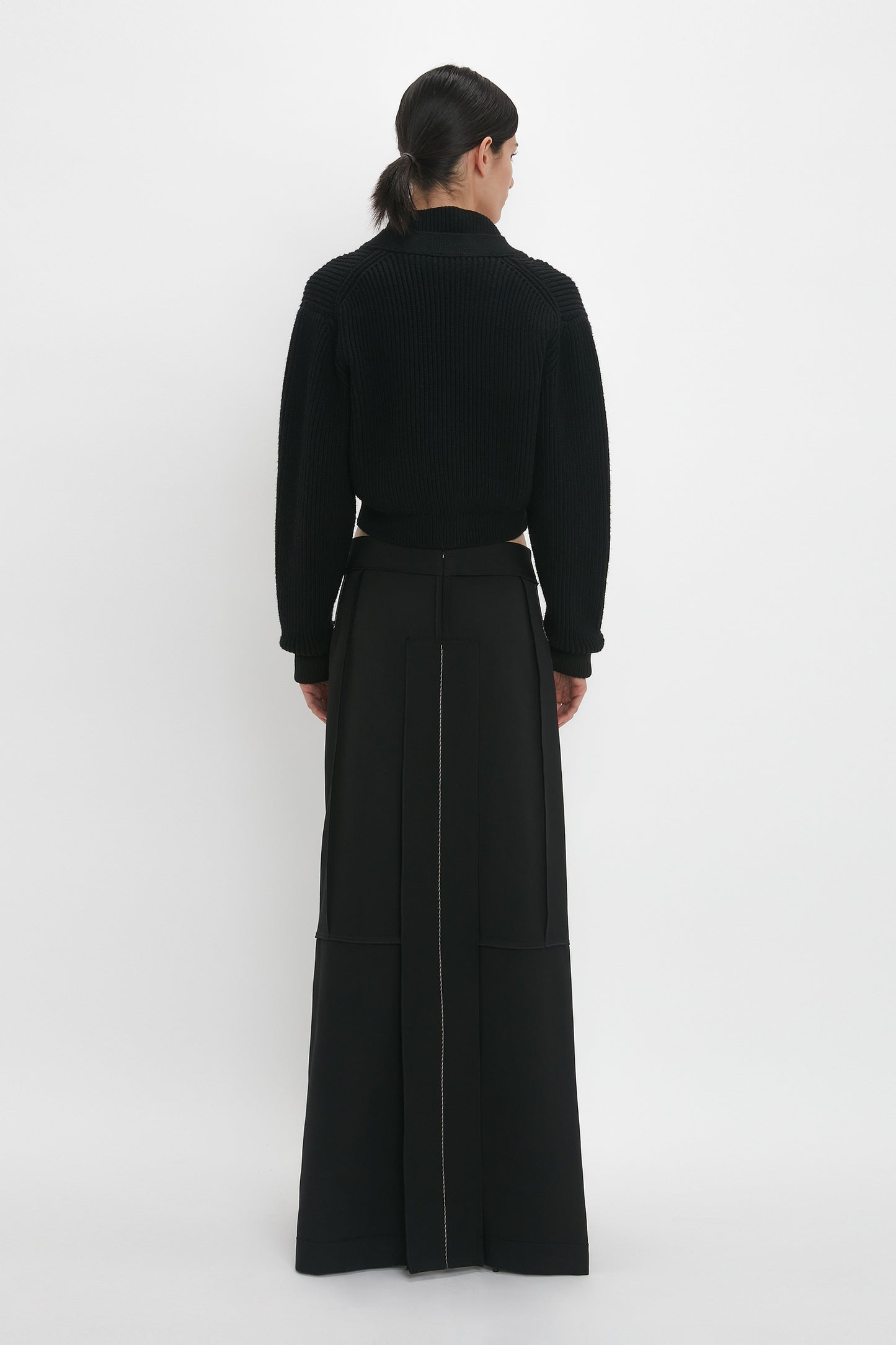 A person stands facing away in a studio setting, wearing a black long-sleeve Cropped V-Neck Cardigan In Black and a long black skirt with seam detailing down the back. The outfit showcases Victoria Beckham monogram elegance and compact ribbed knit sophistication.