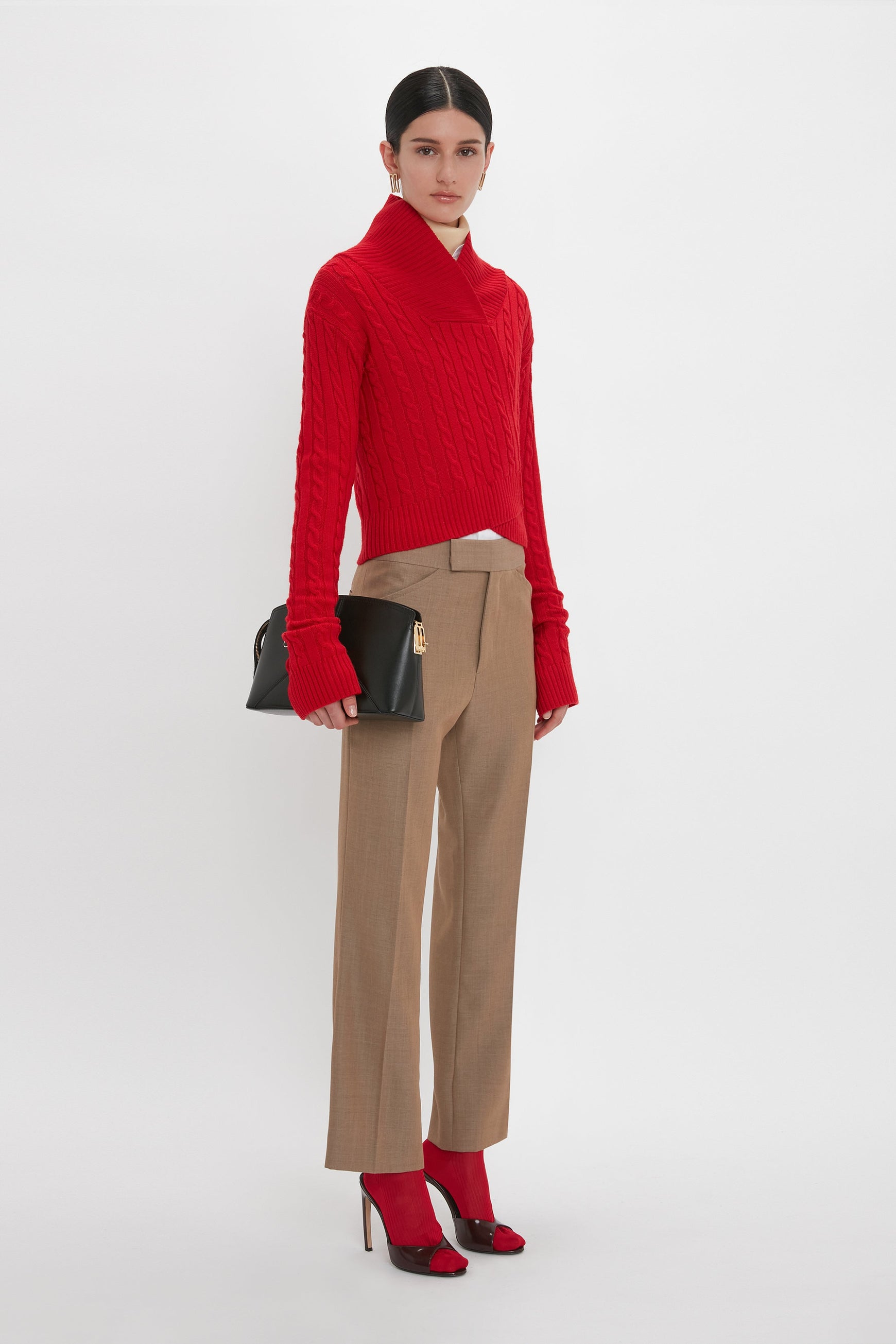 A woman stands against a plain white background wearing a red turtleneck sweater, Victoria Beckham's Wide Cropped Flare Trouser In Tobacco with a contemporary kick hem, red high heels, and holding a black handbag.