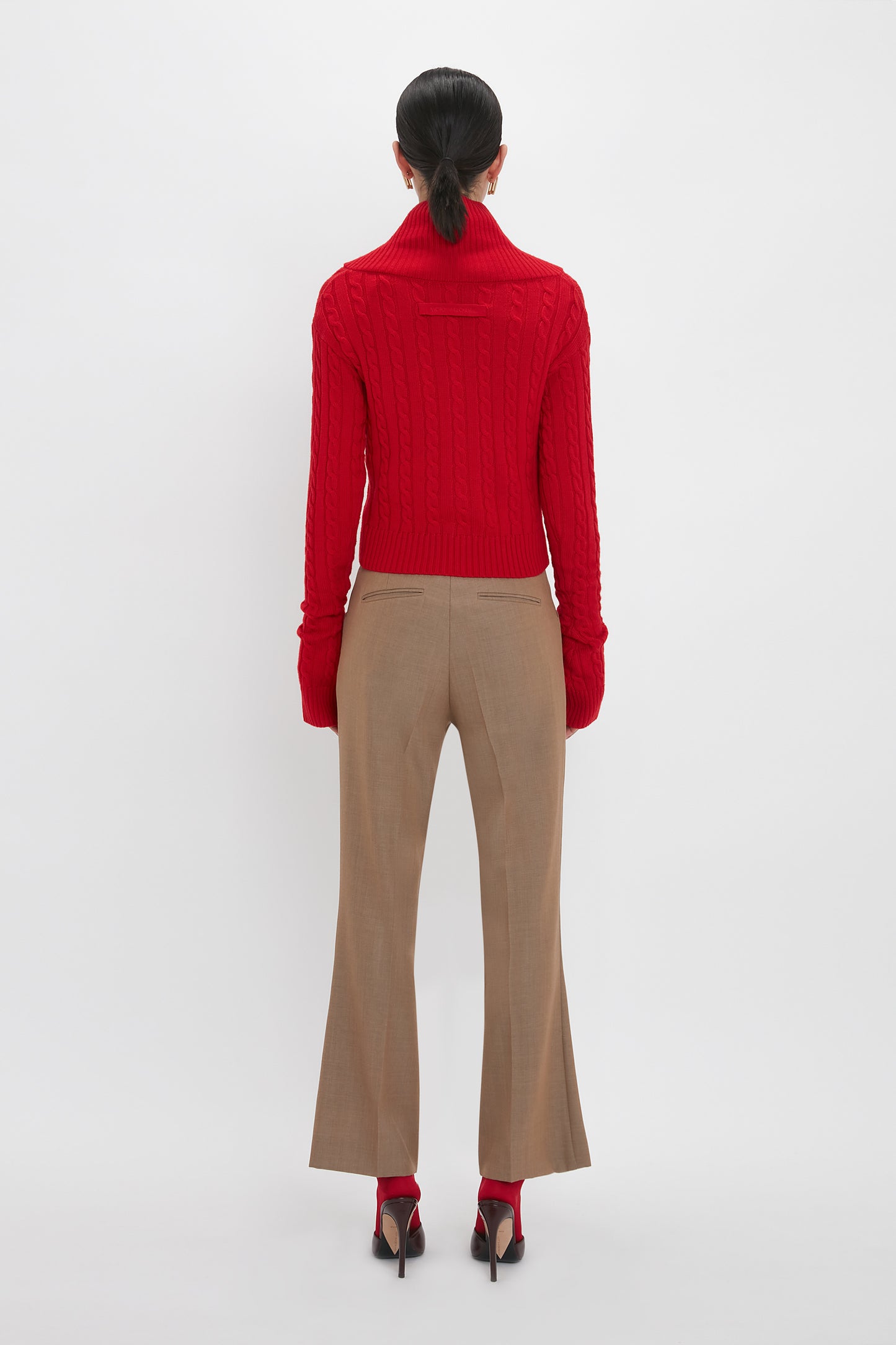 A person stands with their back to the camera, wearing a Wrap Detail Jumper In Red from the Victoria Beckham brand, beige pants, and red shoes with heels. Their hair is tied back in a low ponytail. The luxury knitwear piece contrasts beautifully against the plain white background.