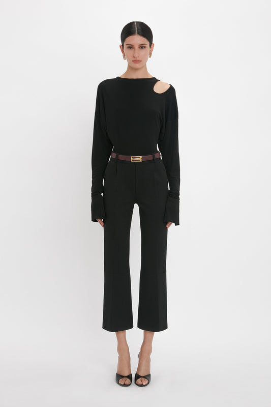 Person in a black outfit stands against a white background. They wear an asymmetrical Twist Detail Jersey Top In Black by Victoria Beckham with cut-out teardrops on one shoulder, black pants, open-toe heels, and a belt.