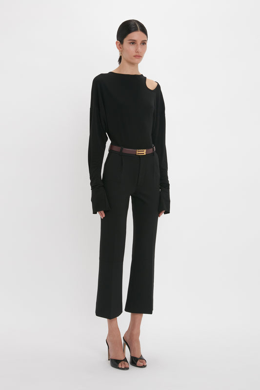 A person stands against a white background wearing a black Victoria Beckham Twist Detail Jersey Top In Black with a shoulder cutout, black pants, a brown belt with a gold buckle, and black high-heeled sandals.