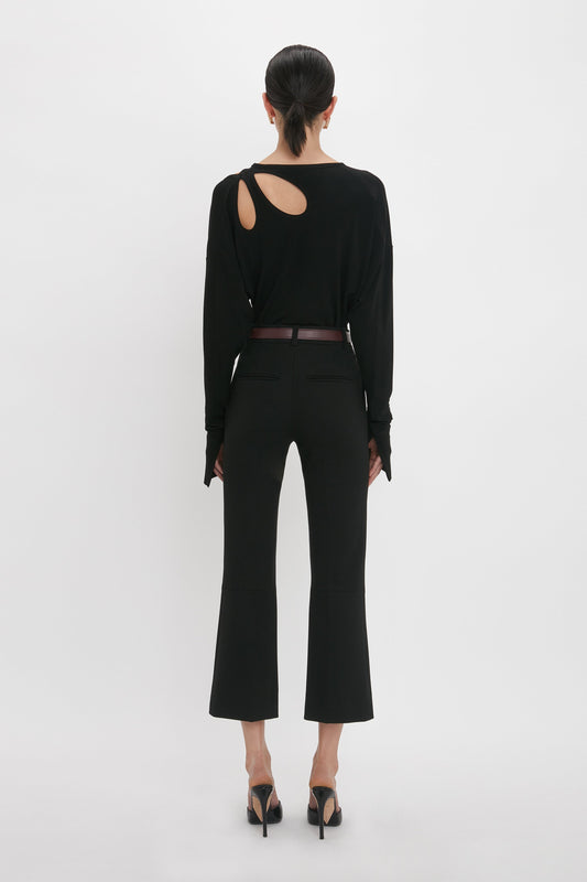 A person with dark hair stands facing away. They wear a black long-sleeve Twist Detail Jersey Top In Black by Victoria Beckham featuring a cutout on the right shoulder, black pants, and black high heels.