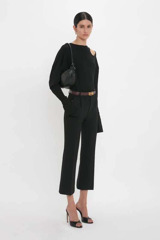 A person dressed in a sleek black outfit, featuring a Twist Detail Jersey Top In Black by Victoria Beckham, fitted pants, and high-heeled sandals, holding a black handbag, stands against a plain white background.