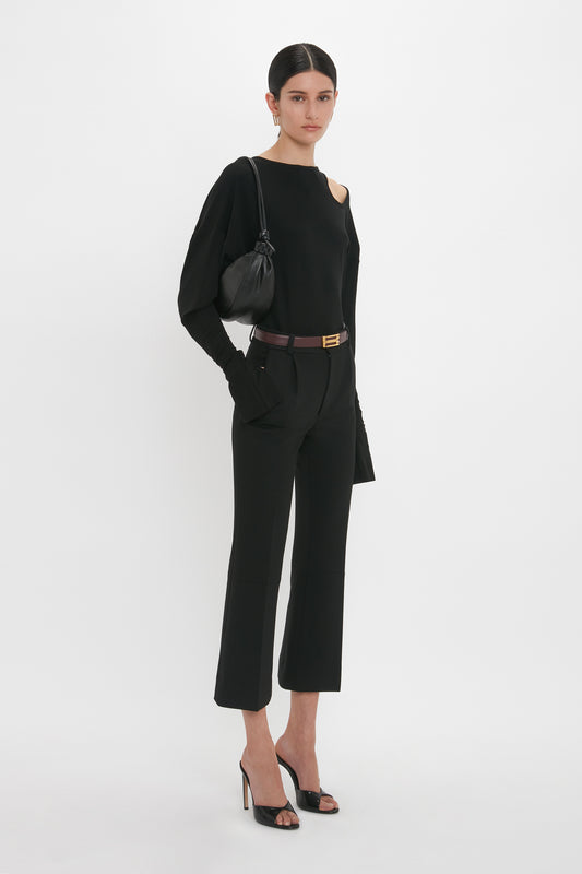 A person stands against a white background, wearing a black outfit with a long-sleeve top, cropped pants, and seductive Classic Mule In Black Calf Leather by Victoria Beckham, holding a black purse with a shoulder strap.