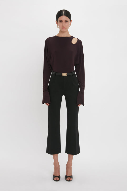A person stands against a white background, wearing a dark long-sleeve Twist Detail Jersey Top In Deep Mahogany by Victoria Beckham with a shoulder cutout, black high-waisted pants, and black open-toe heels, exuding casual sophistication.