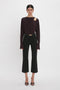 A person stands against a white background, wearing a dark long-sleeve Twist Detail Jersey Top In Deep Mahogany by Victoria Beckham with a shoulder cutout, black high-waisted pants, and black open-toe heels, exuding casual sophistication.