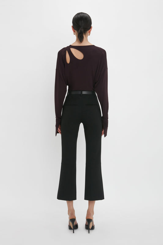 A person stands facing away, exuding casual sophistication in the Twist Detail Jersey Top In Deep Mahogany by Victoria Beckham, black high-waist pants, and black high-heeled shoes.