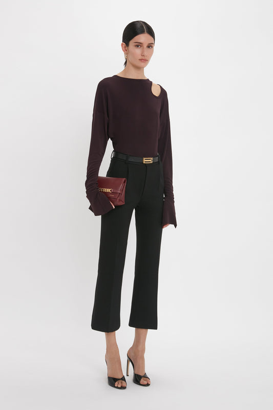 A person stands against a plain background wearing a black Victoria Beckham Twist Detail Jersey Top In Deep Mahogany with a shoulder cutout, black pants, and black heels, exuding casual sophistication while holding a small Deep Mahogany clutch.
