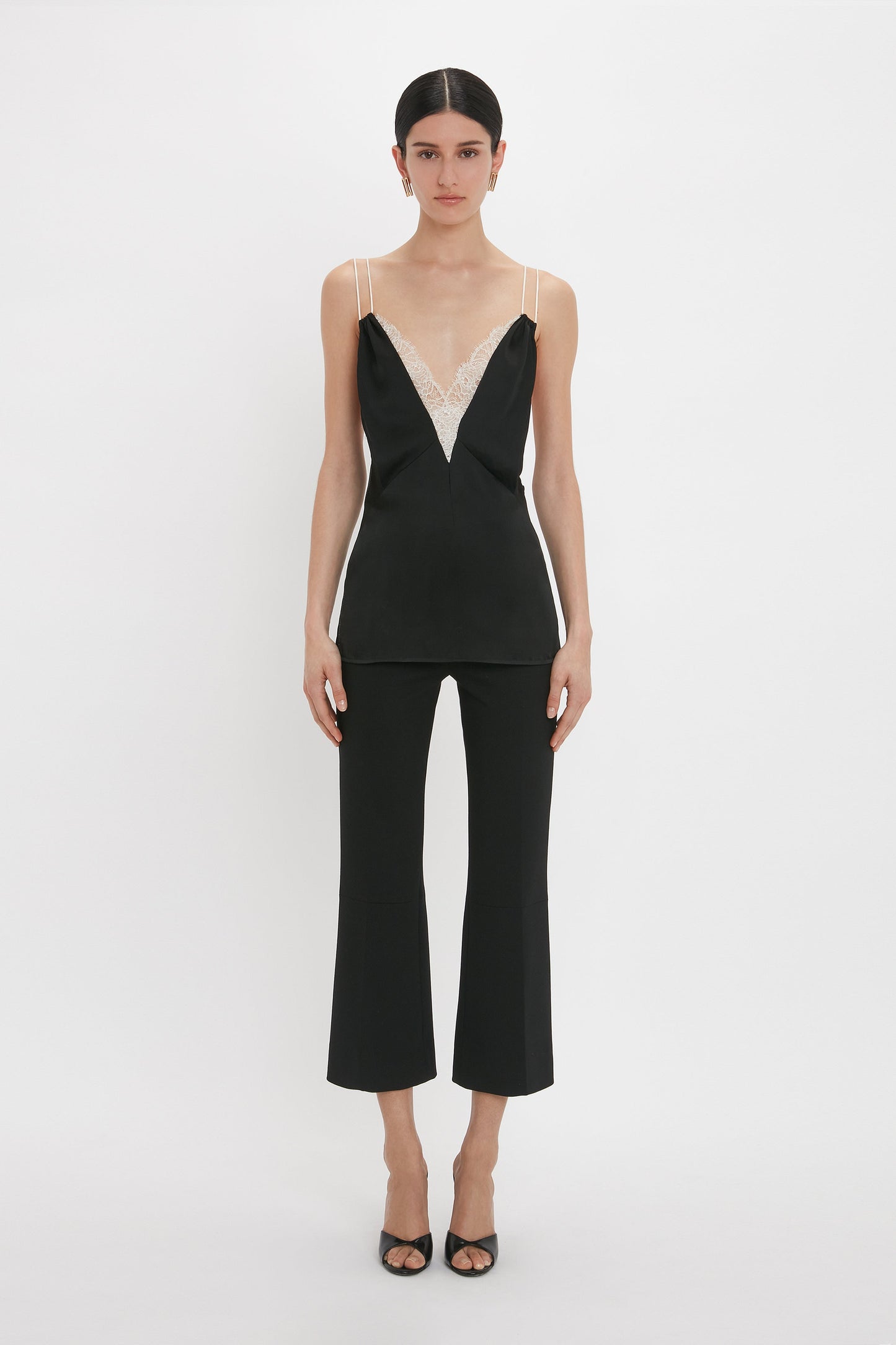 A person stands facing forward wearing the Victoria Beckham Lace Detail Cami Top In Black with a deep-V neckline and matching black pants with flared legs.