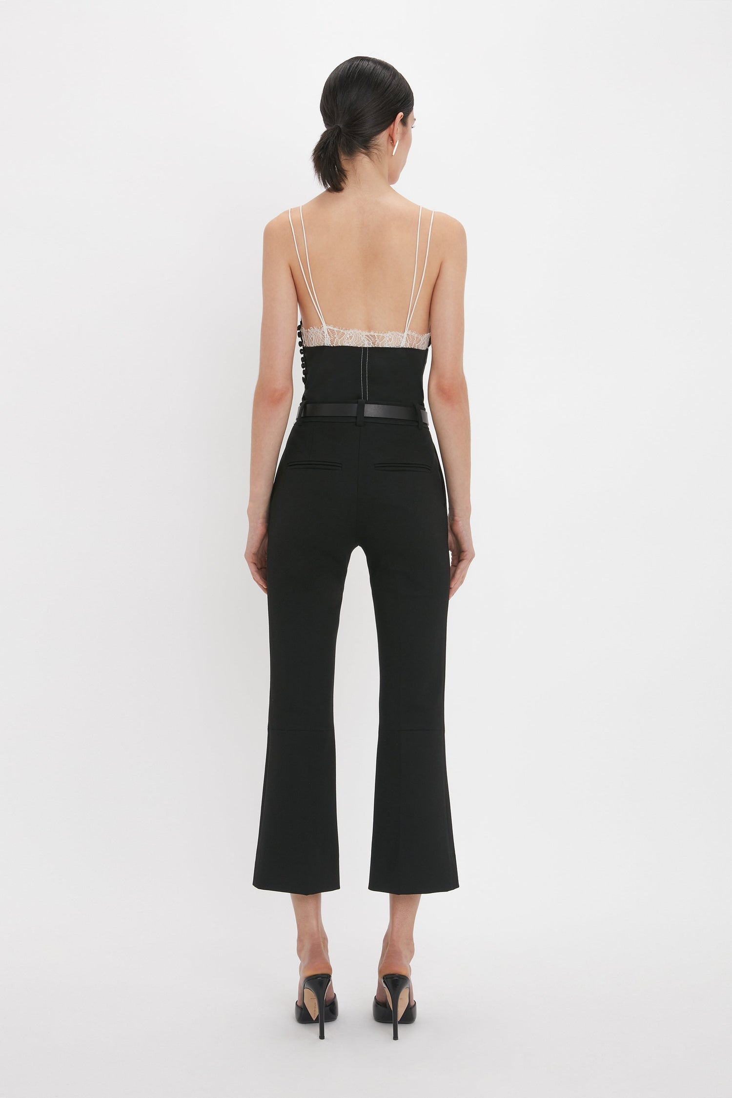 A person with dark hair in a ponytail is wearing a Victoria Beckham Lace Detail Cami Top In Black, featuring a deep-V neckline and black high-heeled shoes, seen from the back against a plain white background.