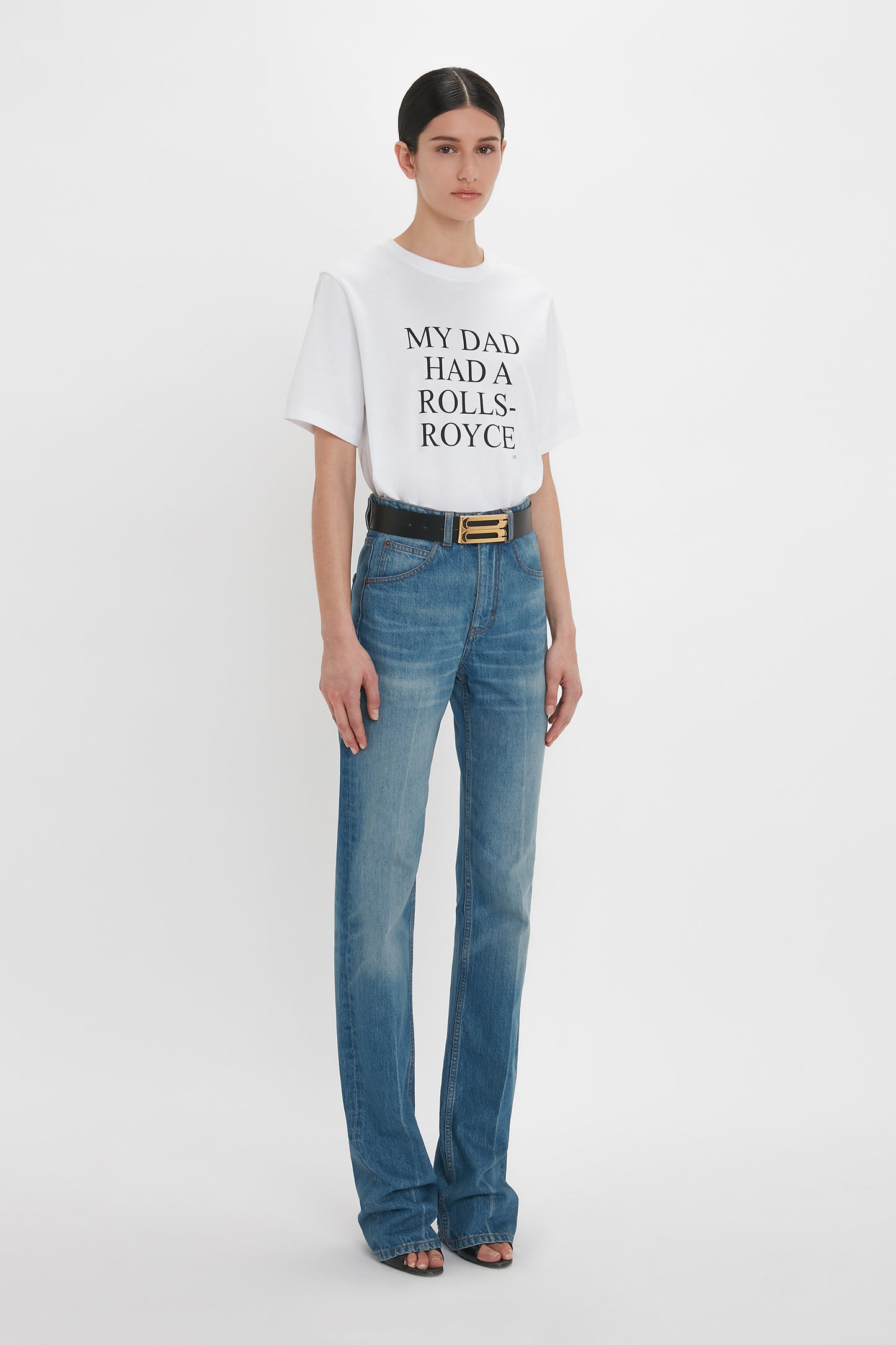 A person stands against a plain white background wearing a white t-shirt with the text "MY DAD HAD A ROLLS-ROYCE," high waistline blue jeans in distressed Julia Jean In Broken Vintage Wash by Victoria Beckham, and black open-toe sandals.