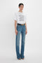 A person stands against a plain white background wearing a white t-shirt with the text "MY DAD HAD A ROLLS-ROYCE," high waistline blue jeans in distressed Julia Jean In Broken Vintage Wash by Victoria Beckham, and black open-toe sandals.