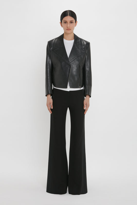 Person standing against a white background, wearing a Victoria Beckham Tailored Leather Biker Jacket In Black over a white shirt and black wide-legged trousers.