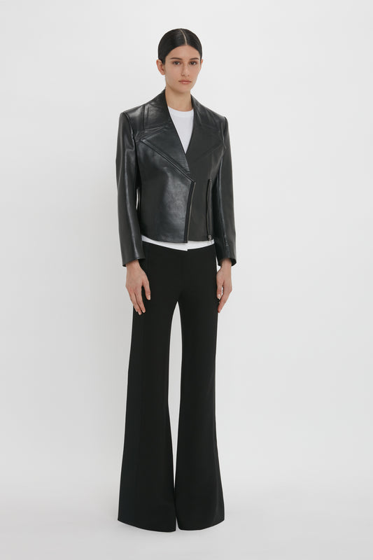 A person stands wearing a Victoria Beckham Tailored Leather Biker Jacket In Black over a white shirt and black wide-leg pants, set against a plain white background, exuding a modern classic vibe.