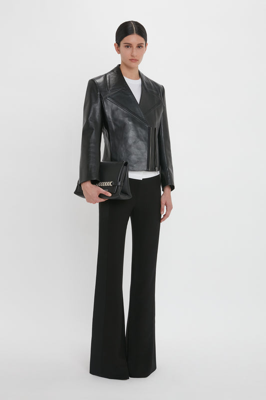 A person stands against a white background, wearing a Tailored Leather Biker Jacket In Black by Victoria Beckham, a white shirt, black wide-leg pants, and holding a black clutch.