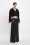 A person stands against a plain background, wearing a black long-sleeve cropped top and a Deconstructed Floor-Length Skirt In Black by Victoria Beckham with a front slit. They hold a black clutch and have their hair pulled back, creating an elongated silhouette.