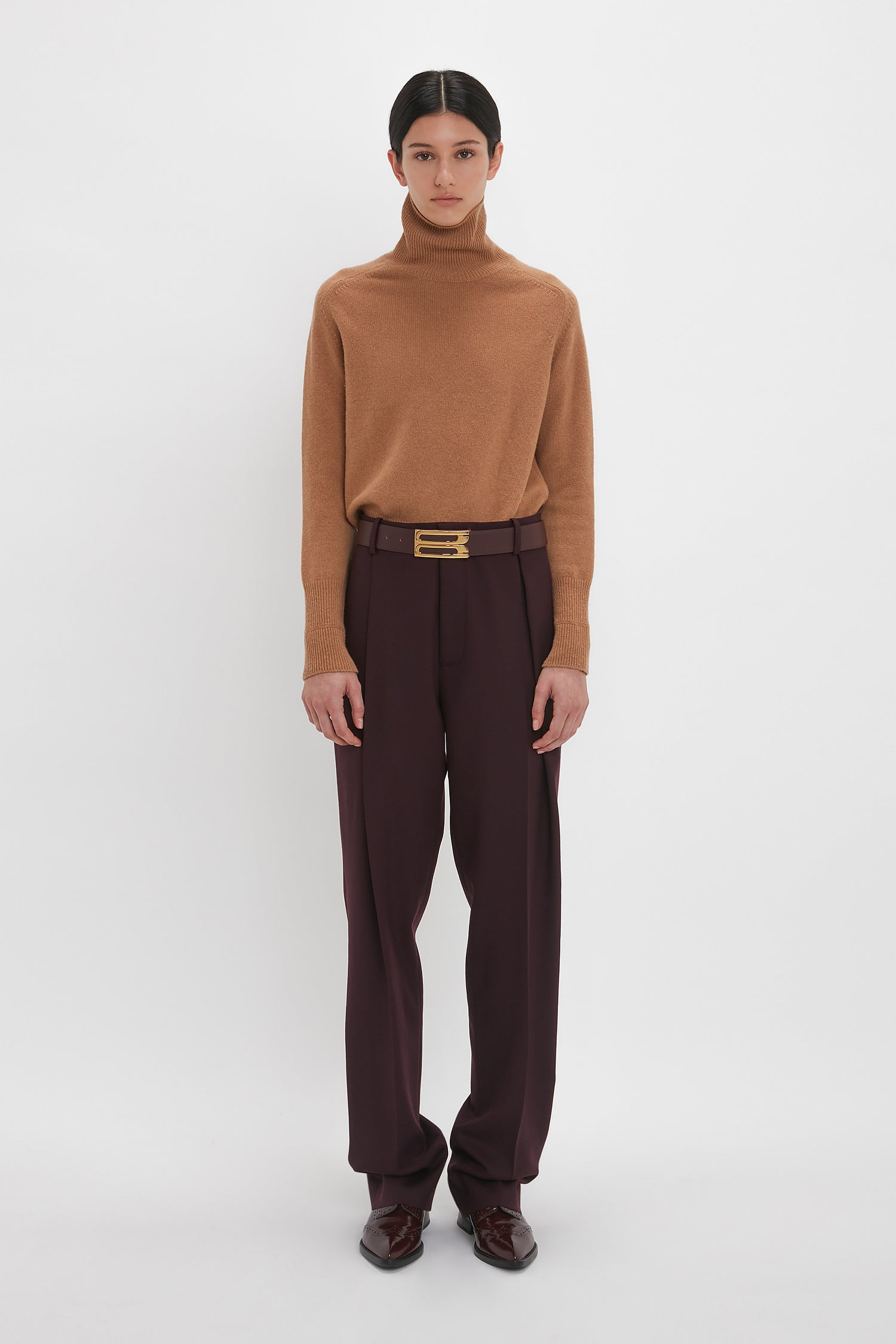 Person standing against a white background, wearing a luxurious Polo Neck Jumper In Tobacco by Victoria Beckham, dark maroon pants with a belt, and brown shoes.