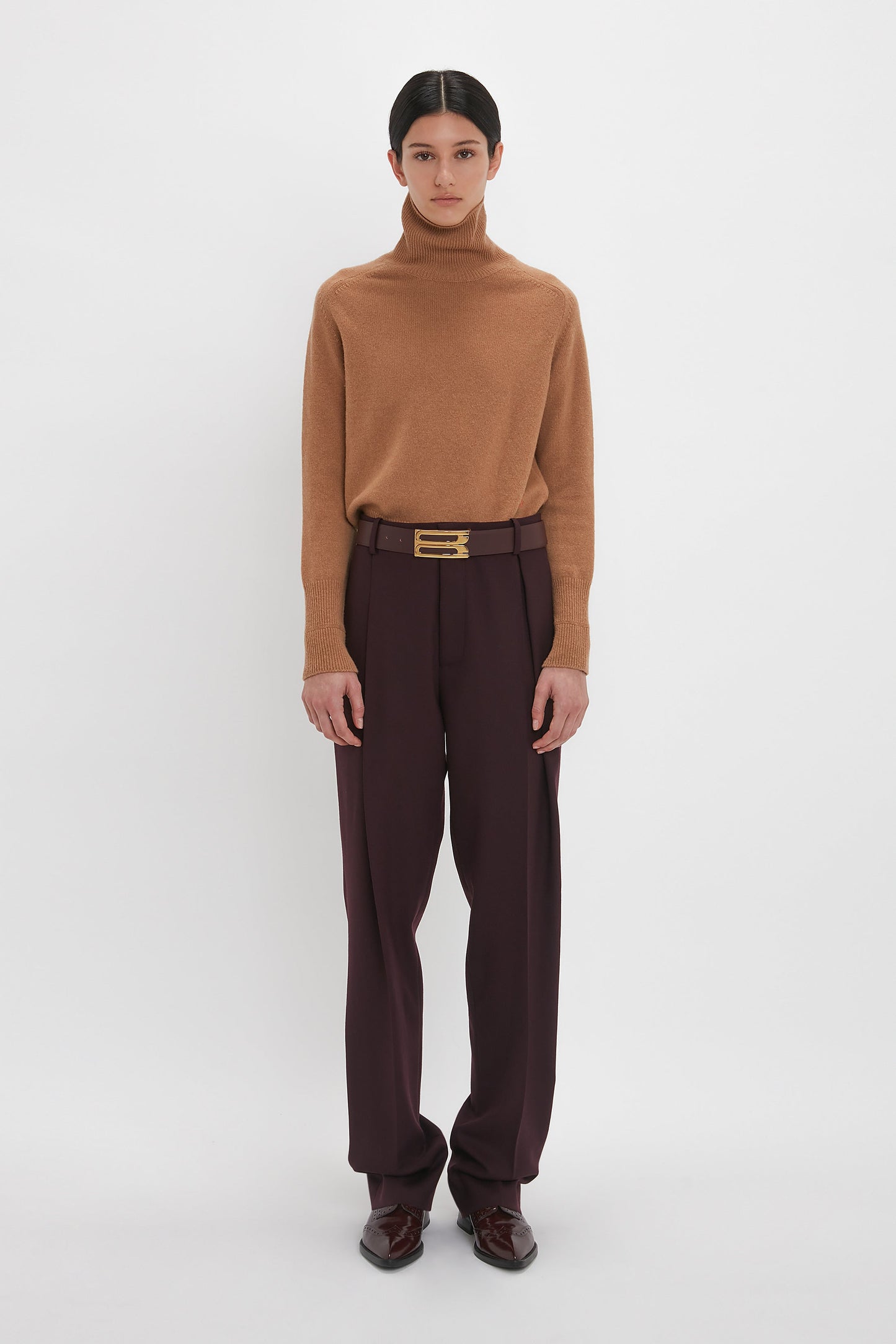 A person with dark hair wearing a brown turtleneck sweater, Victoria Beckham Asymmetric Chino Trouser In Deep Mahogany with a narrow leg silhouette, and dark shoes, standing against a plain white background.