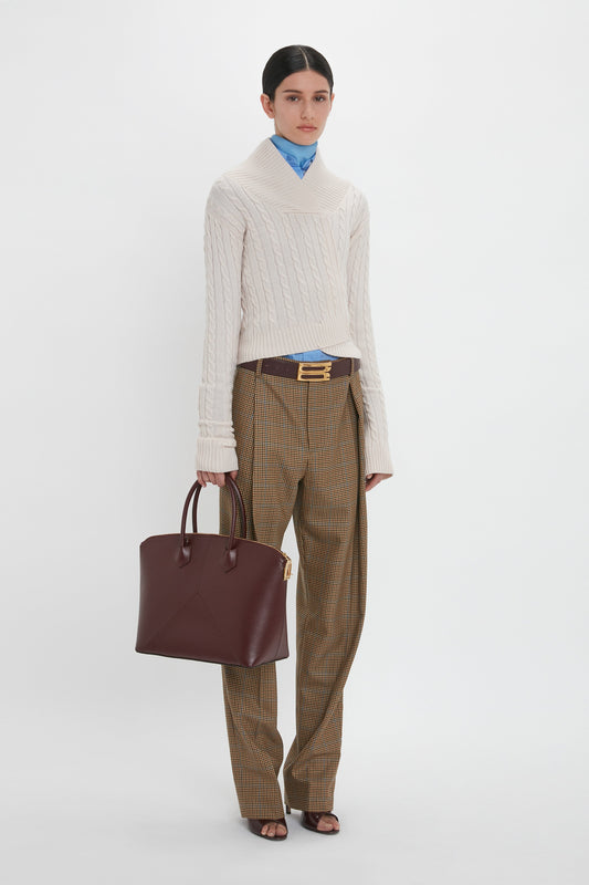 Person in a cream sweater and brown plaid pants holding a Victoria Beckham Victoria Bag In Burgundy Leather with a padlock closure against a plain white background.