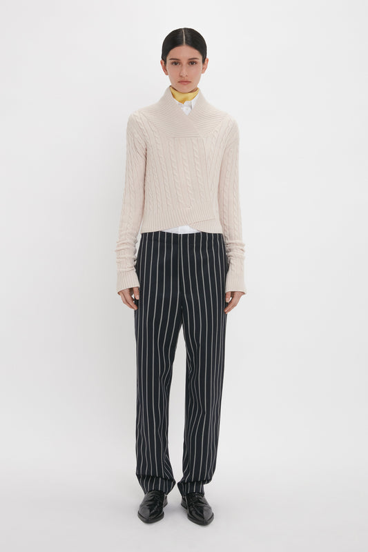Person stands against a plain background, wearing a light-colored knit sweater over a shirt with a yellow collar, and Tapered Leg Trouser In Midnight-White by Victoria Beckham. They have dark hair pulled back and wear black shoes.