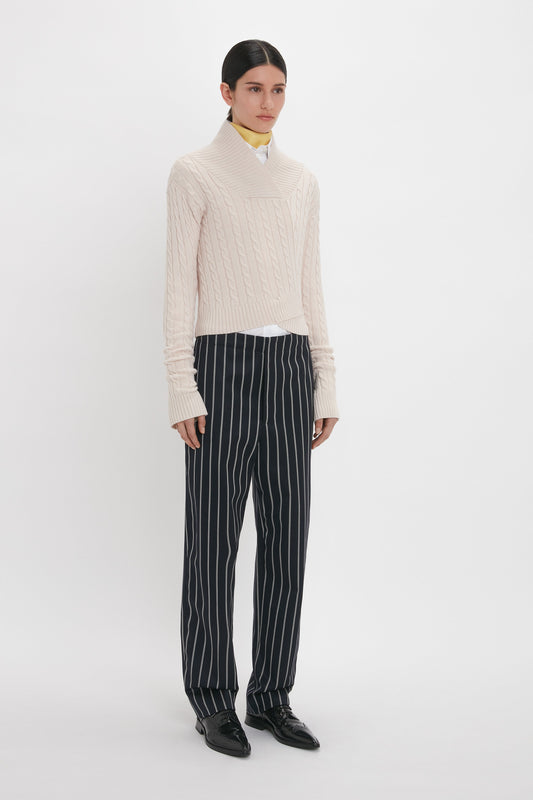 A person stands against a plain background wearing a cream cable-knit sweater and Victoria Beckham's Tapered Leg Trouser In Midnight-White, paired with black shoes.