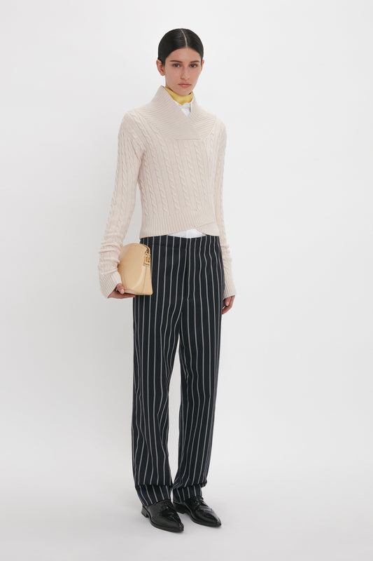 A person stands against a white background, wearing a cream cable-knit sweater, Tapered Leg Trouser In Midnight-White by Victoria Beckham, and holding a small beige handbag.