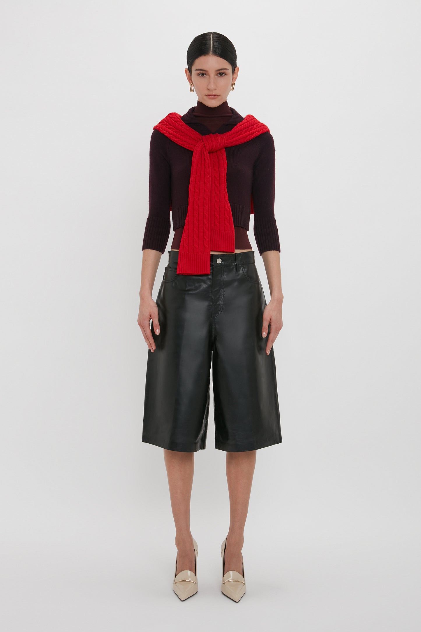 A person stands against a white background wearing a red sweater around their neck, a dark shirt, Victoria Beckham's Leather Bermuda Short In Black, and beige pointed shoes.