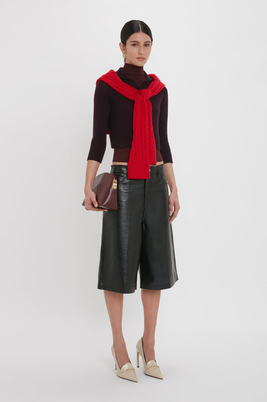 A woman stands against a plain background wearing a dark top with a red sweater draped over her shoulders, Victoria Beckham Leather Bermuda Short In Black, beige high heels, and holding a burgundy clutch bag.