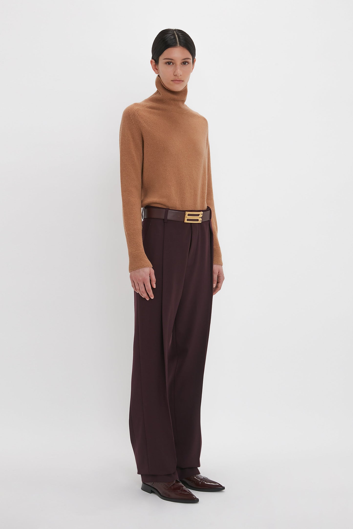 A person with short hair stands wearing a brown turtleneck sweater, Victoria Beckham Asymmetric Chino Trouser in Deep Mahogany, a black belt with a gold buckle, and brown shoes, against a plain white background.