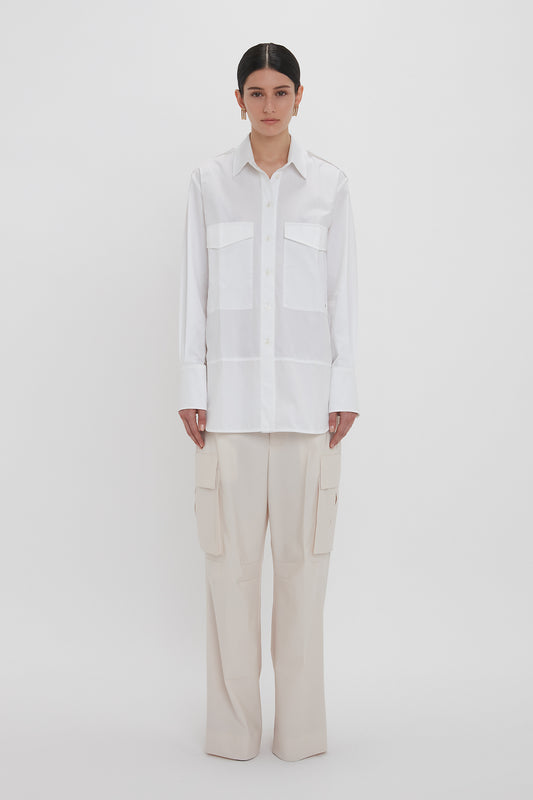 Person standing against a white background, wearing an **Oversized Pocket Shirt In White** crafted from organic cotton with two chest pockets and beige cargo pants, exuding modern sophistication. The shirt is by **Victoria Beckham**.