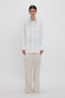 Person standing against a white background, wearing an **Oversized Pocket Shirt In White** crafted from organic cotton with two chest pockets and beige cargo pants, exuding modern sophistication. The shirt is by **Victoria Beckham**.