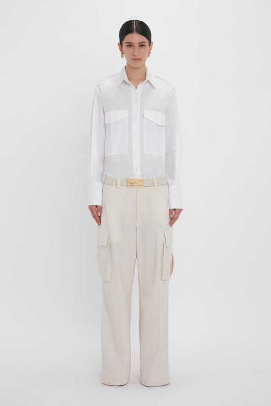 A person with dark hair is wearing a white collared shirt and cream-colored, 100% cotton Relaxed Cargo Trouser In Bone by Victoria Beckham, standing against a plain white background. The military-inspired aesthetic adds a touch of rugged elegance to the outfit.