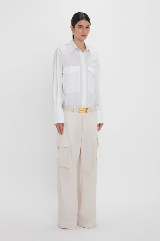 Person standing against a white background, wearing the Oversized Pocket Shirt In White by Victoria Beckham made from organic cotton and beige wide-leg cargo pants with a belt, showcasing modern sophistication.