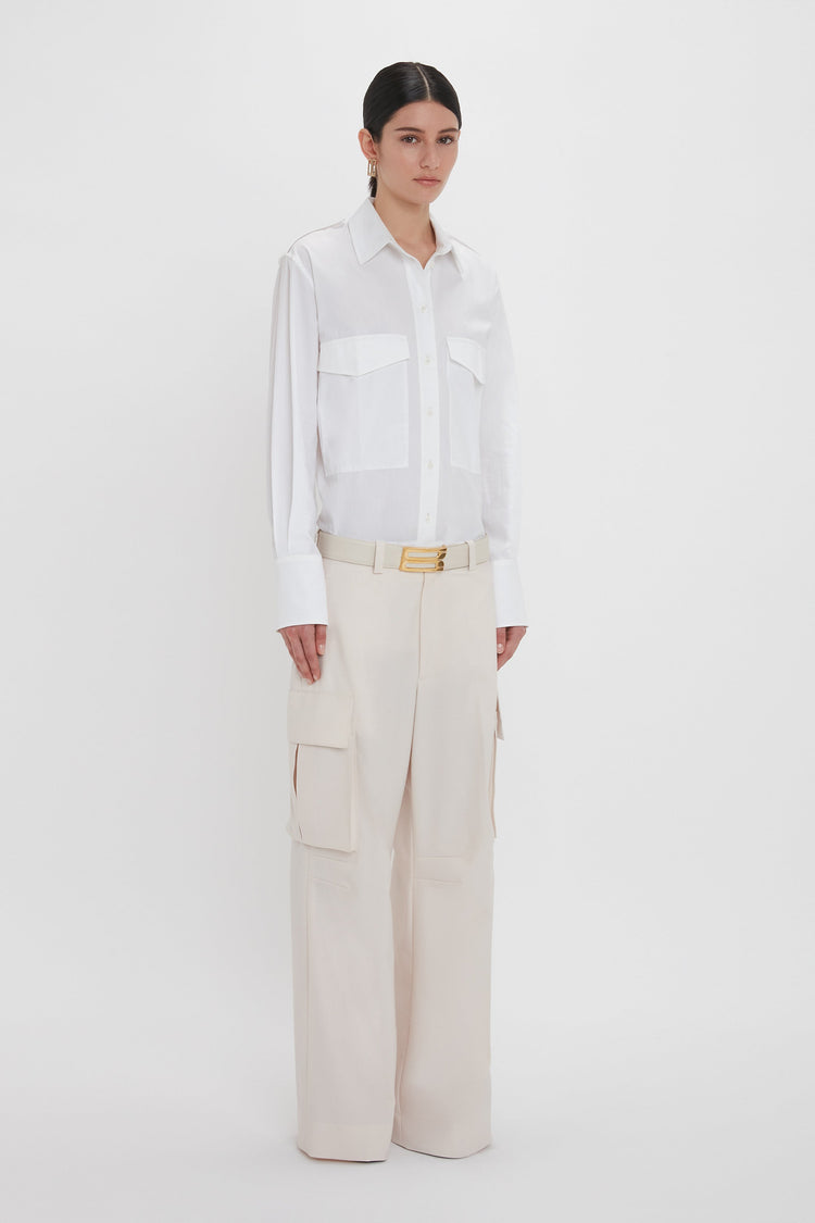 Person standing against a white background, wearing a white button-up shirt and Victoria Beckham's Relaxed Cargo Trouser In Bone with pockets, accessorized with a belt. The 100% cotton material offers comfort while adding a touch of military-inspired style.