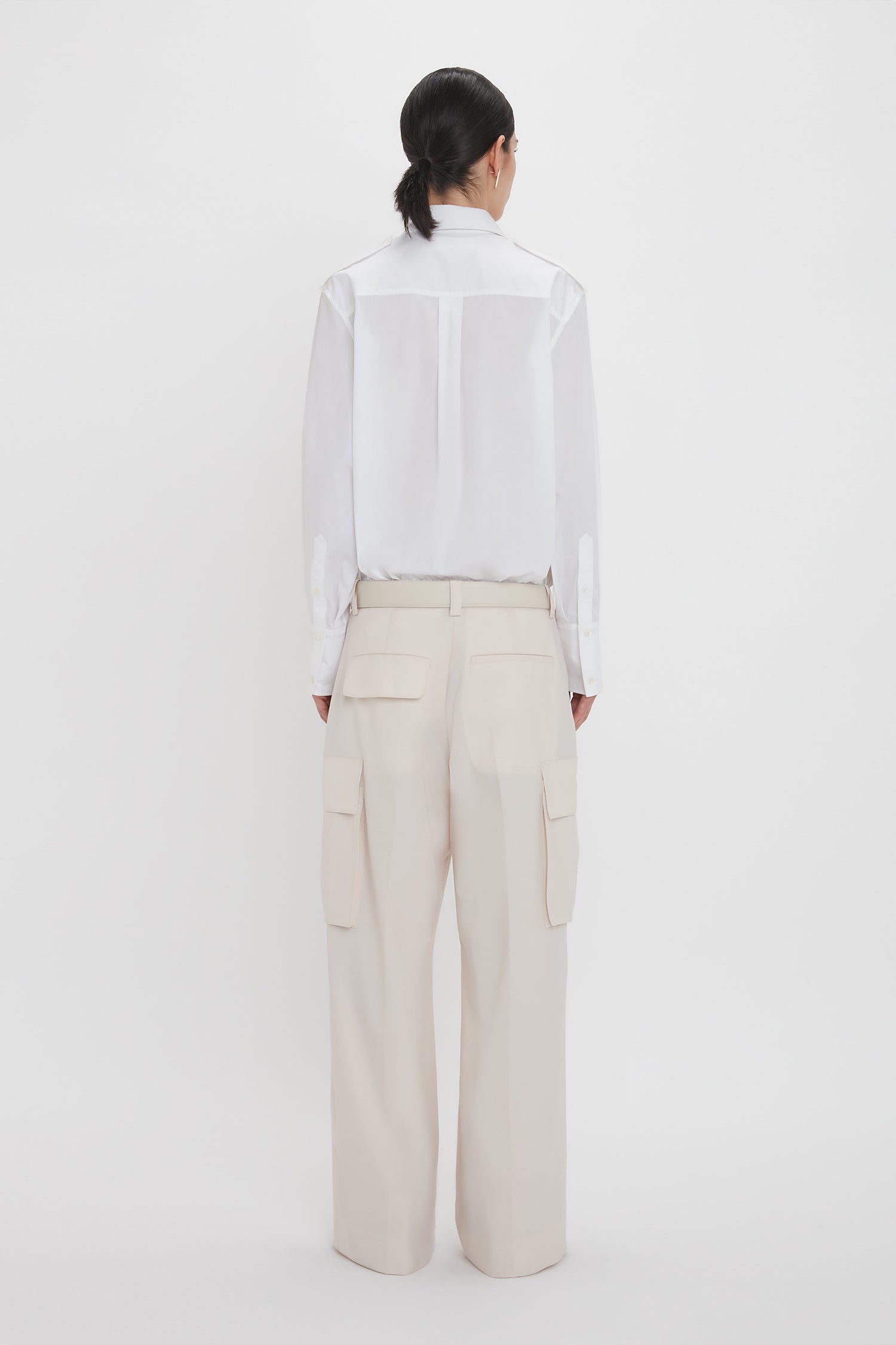 A person with dark hair is standing and facing away, wearing an Oversized Pocket Shirt In White by Victoria Beckham made of organic cotton paired with beige high-waisted pants featuring large pockets, showcasing modern sophistication.