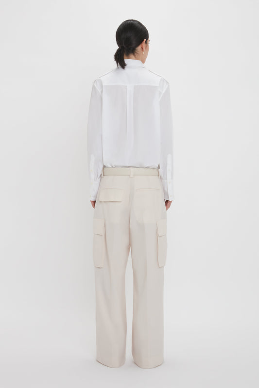 A person with dark hair in a ponytail wearing a white long-sleeve shirt and Victoria Beckham's Relaxed Cargo Trouser In Bone, crafted from 100% cotton, stands facing away against a plain white background.