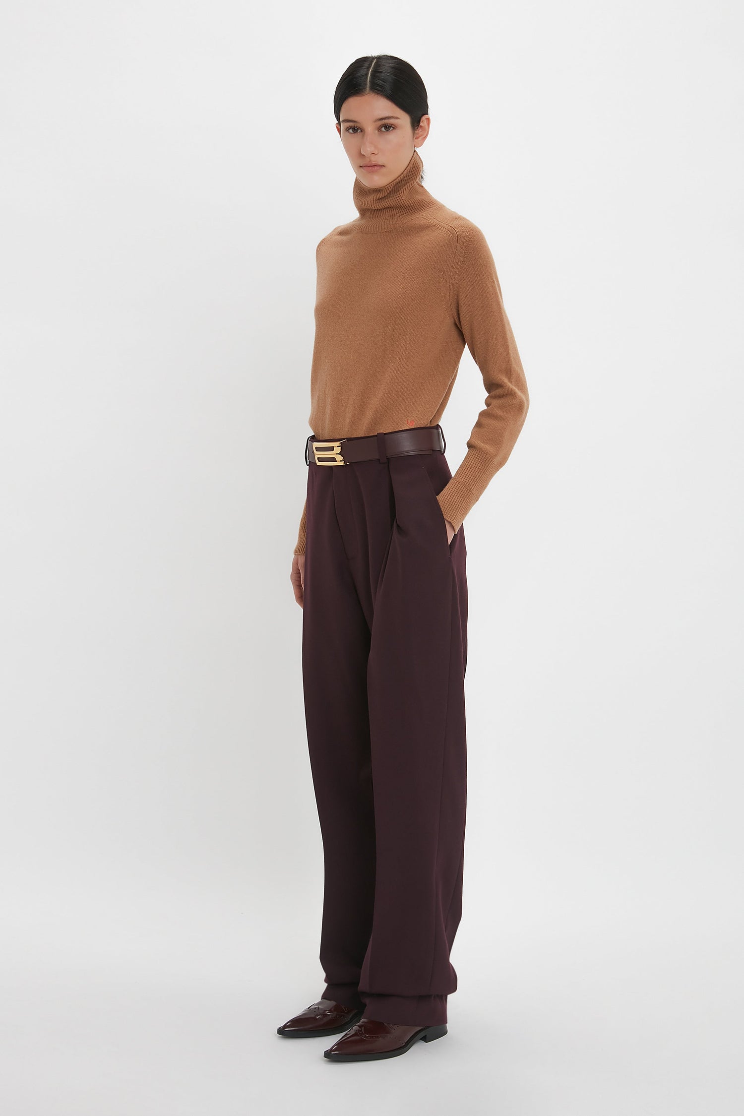 Person standing against a plain white background, wearing a camel-colored turtleneck sweater, Victoria Beckham Asymmetric Chino Trouser In Deep Mahogany with a belt, and dark brown shoes.