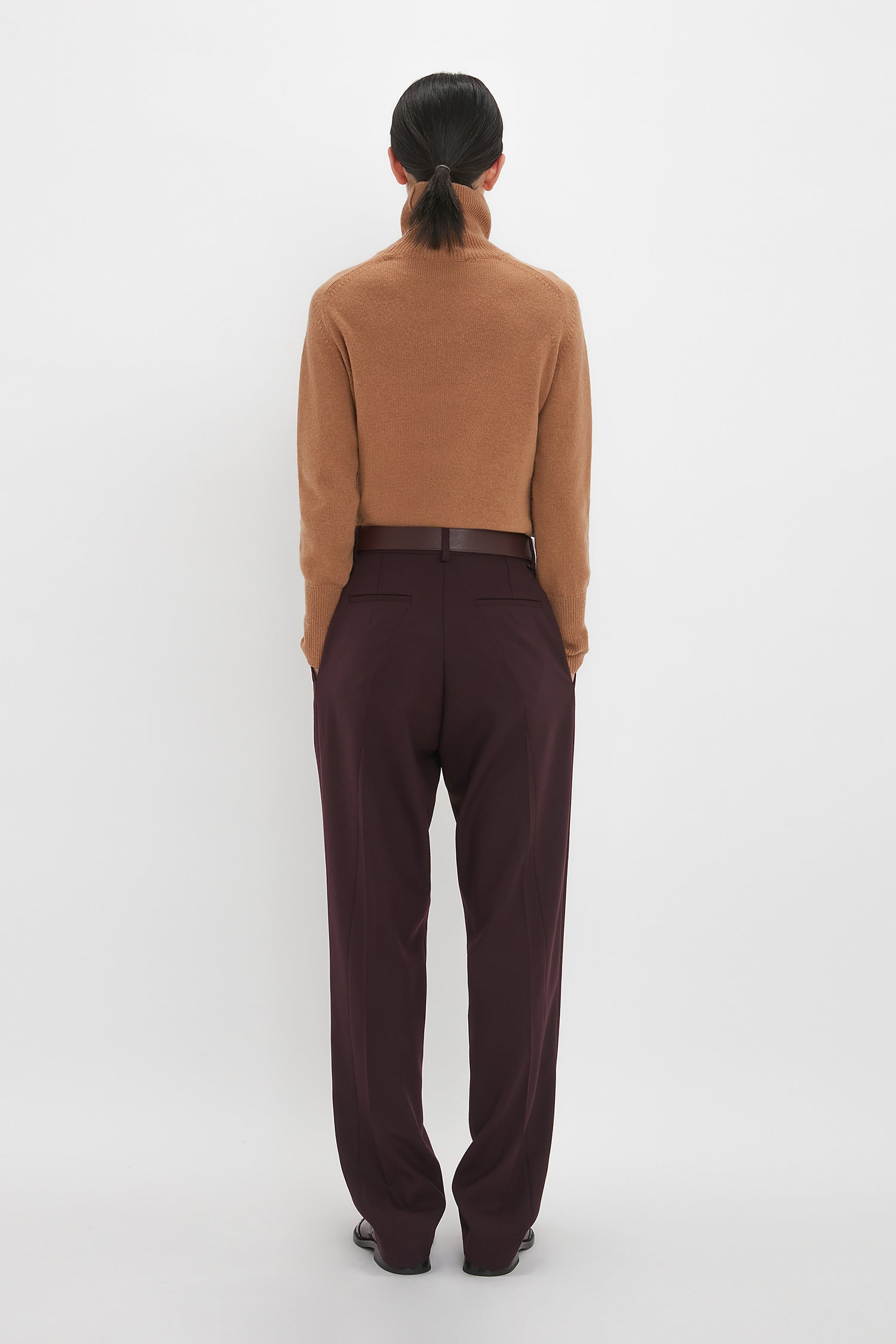 Person standing with back facing camera, wearing a Victoria Beckham Polo Neck Jumper In Tobacco and purple pants against a white background.