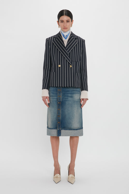 A person stands against a plain background wearing a dark pinstripe blazer, a light blue scarf, the Victoria Beckham Placket Detail Denim Skirt In Heavy Vintage Indigo Wash featuring an asymmetric front placket design, and cream pointed-toe heels.
