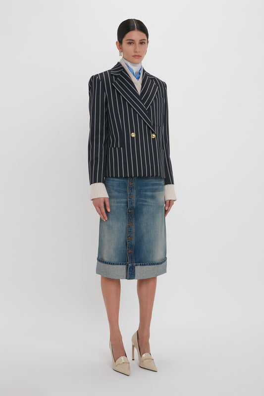 A person stands against a white background, wearing a black and white striped blazer, a light blue collared shirt, beige high heels, and the Victoria Beckham Placket Detail Denim Skirt In Heavy Vintage Indigo Wash. Their dark hair is tied back neatly.