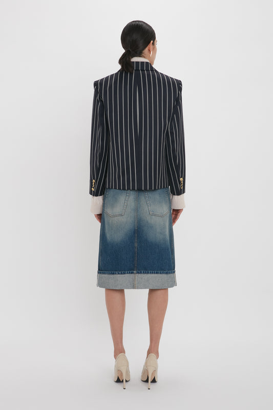 A person stands facing away, wearing a black and white pinstriped blazer, a Placket Detail Denim Skirt In Heavy Vintage Indigo Wash by Victoria Beckham, and beige high heels.