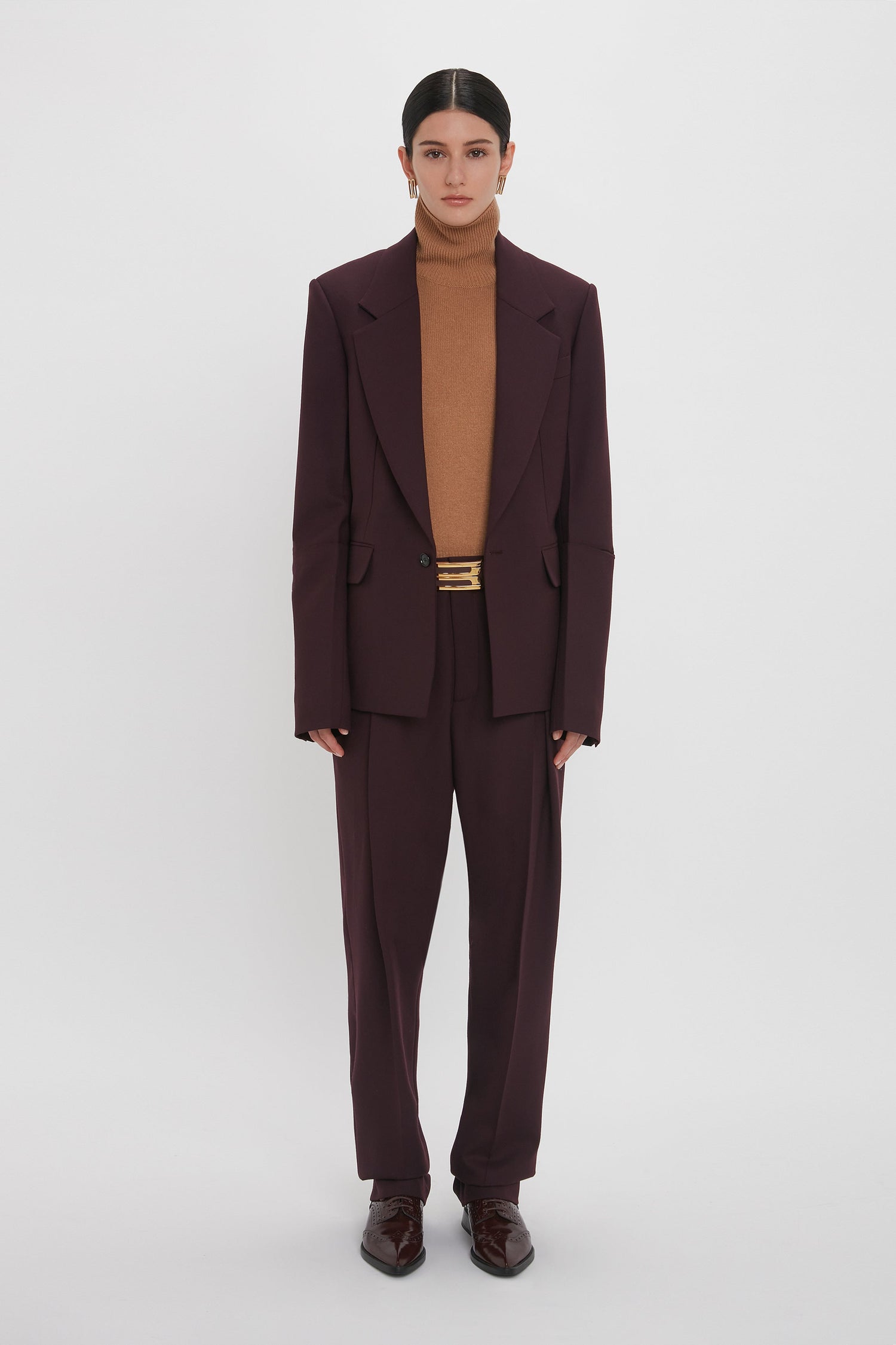 A person stands against a white background wearing a Sleeve Detail Patch Pocket Jacket in Deep Mahogany by Victoria Beckham, a brown turtleneck sweater, and dark shoes.