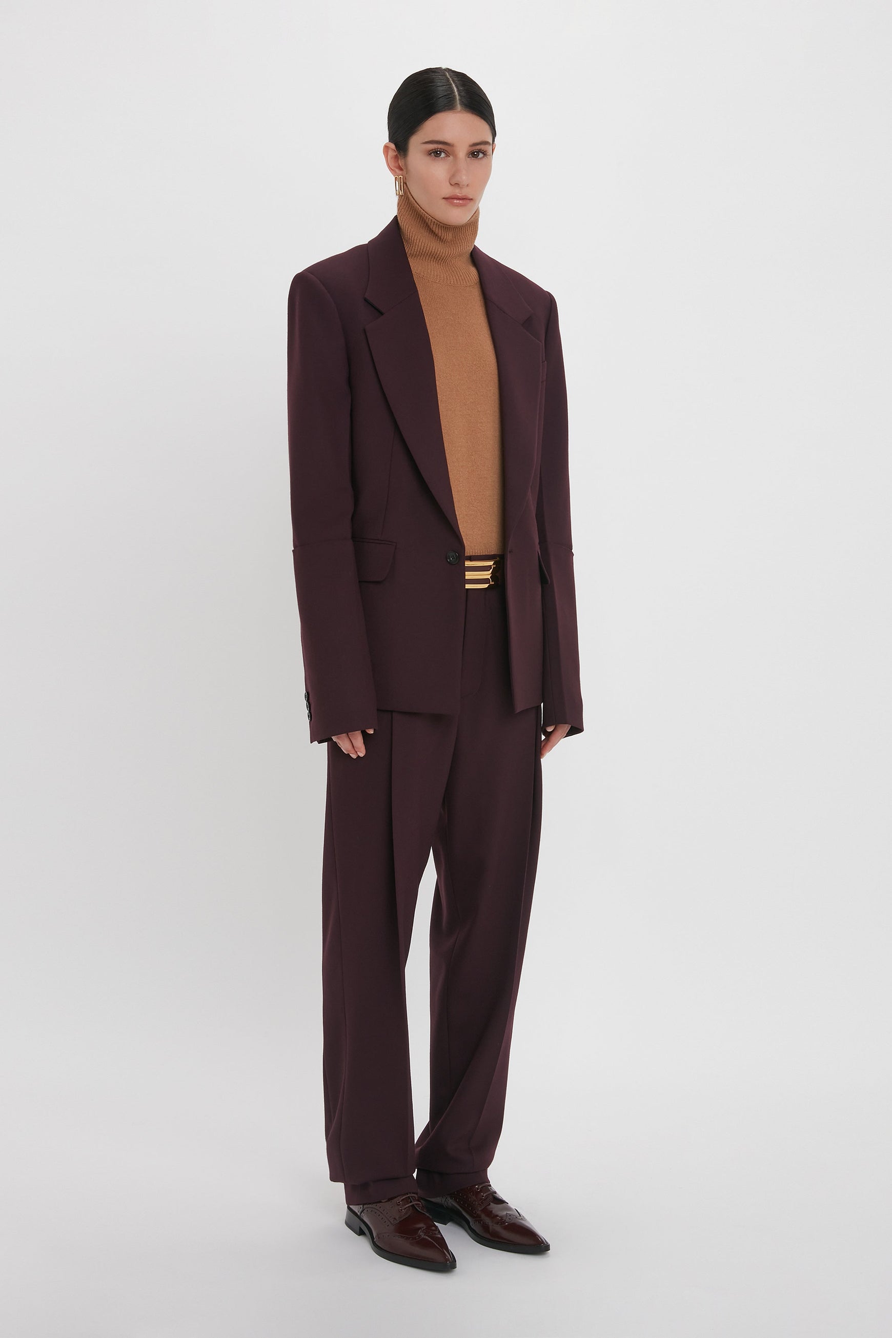 Person wearing a Victoria Beckham Sleeve Detail Patch Pocket Jacket In Deep Mahogany with matching trousers, a tan turtleneck sweater, and dark brown shoes, standing against a plain white background.