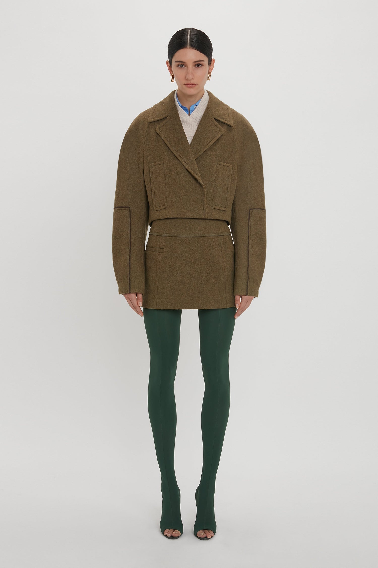 A person stands against a white background wearing a brown oversized jacket, a Tailored Mini Skirt In Khaki by Victoria Beckham, green tights, and open-toe shoes.
