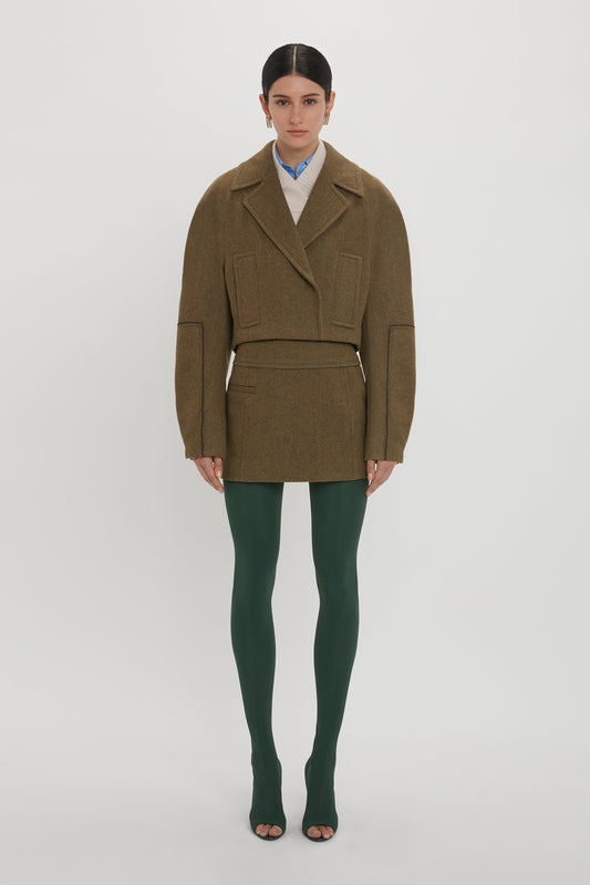 A person stands against a white background wearing a brown oversized jacket, a Tailored Mini Skirt In Khaki by Victoria Beckham, green tights, and open-toe shoes.