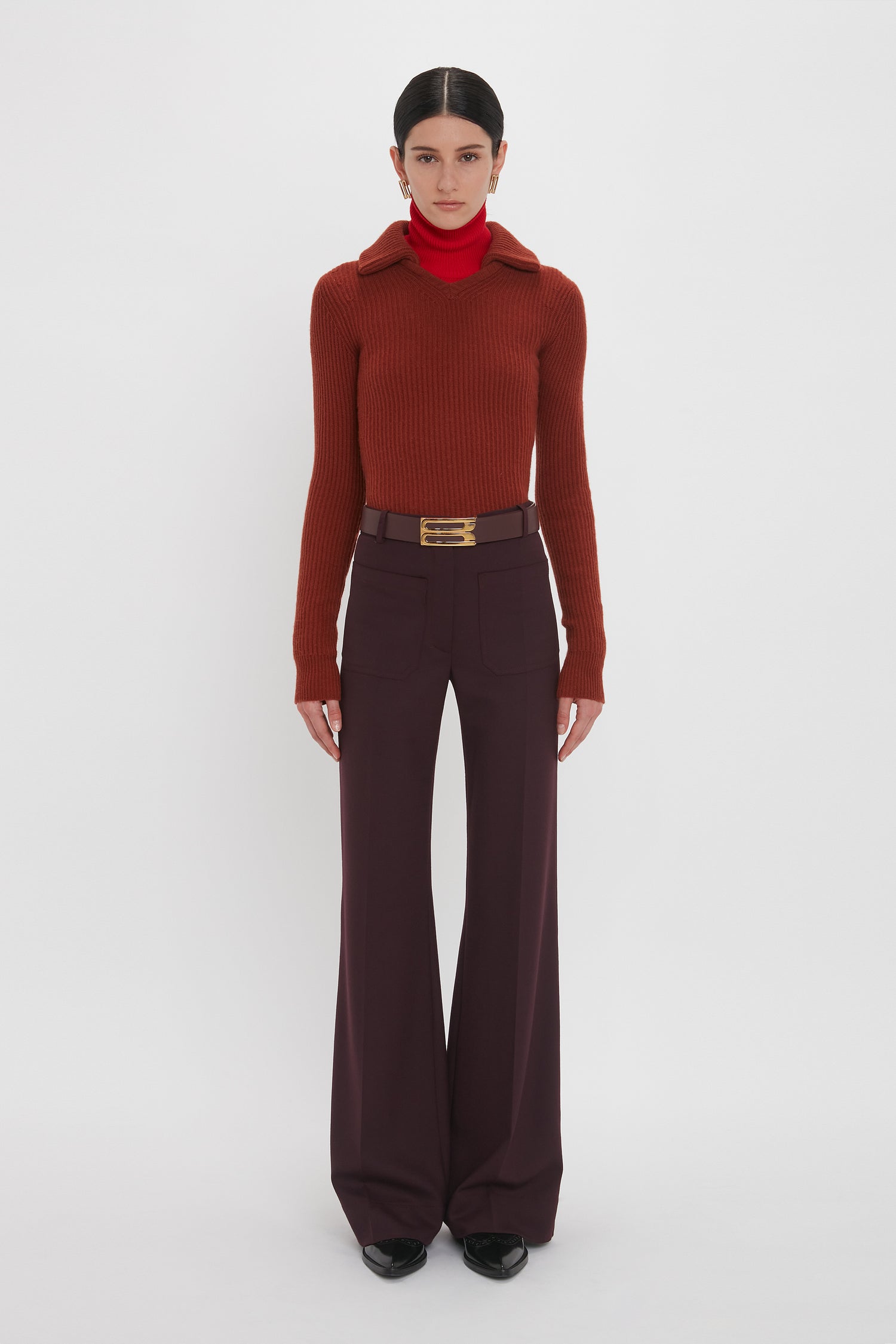 Person wearing a rust-colored Victoria Beckham Double Collared Jumper In Russet, red turtleneck, wide-legged purple pants, and a large belt with a gold buckle, standing against a white background.