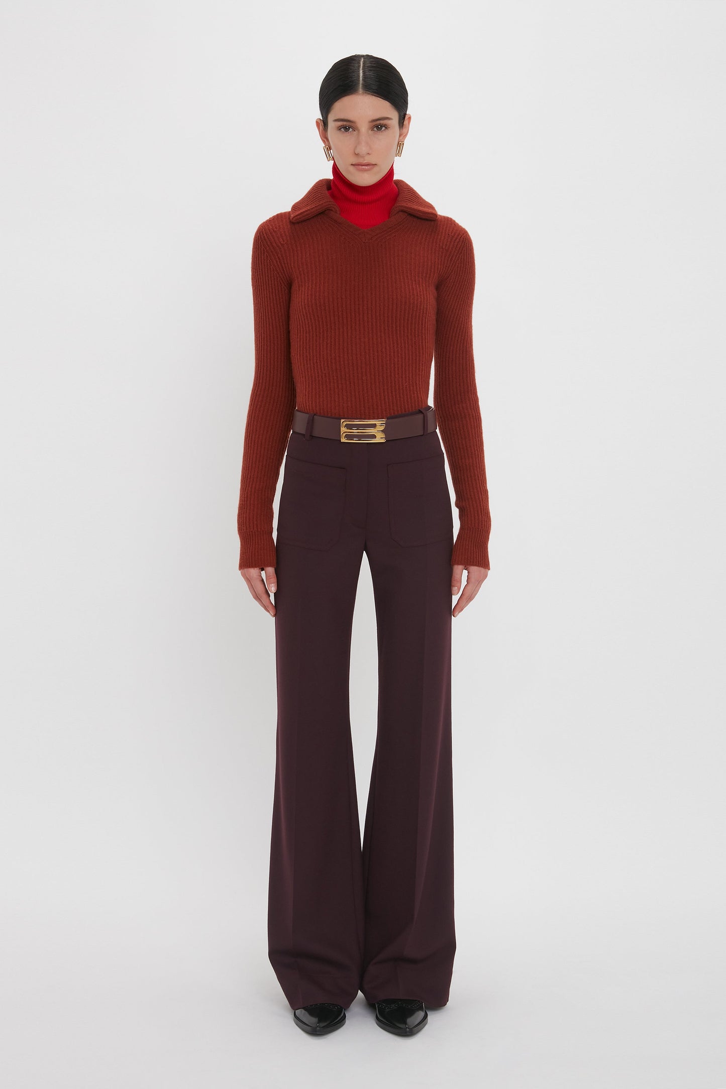 A person stands against a white background, wearing a red turtleneck under a rust-colored sweater made from recycled wool, paired with high-waisted, dark purple Alina Trouser In Deep Mahogany and a belt with a gold buckle. This look echoes the sophisticated style of Victoria Beckham.