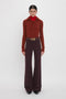 A person stands against a white background, wearing a red turtleneck under a rust-colored sweater made from recycled wool, paired with high-waisted, dark purple Alina Trouser In Deep Mahogany and a belt with a gold buckle. This look echoes the sophisticated style of Victoria Beckham.